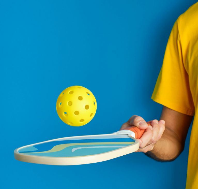 hand with paddle and yellow pickleball ball on blue background